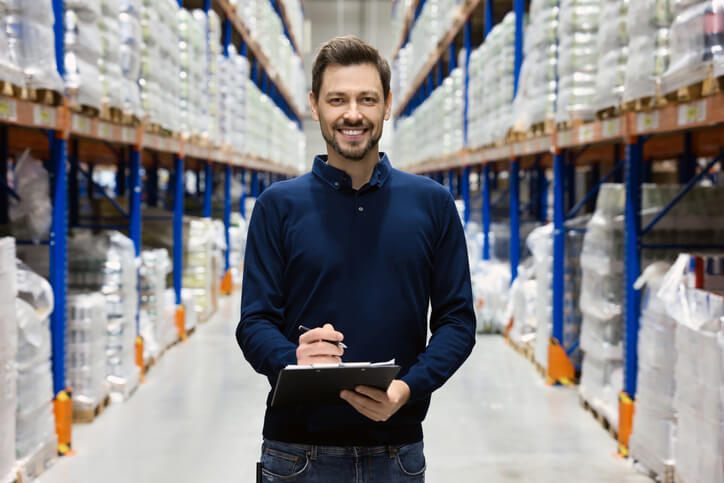 A smiling male sales assistant taking stock of inventory at a warehouse after completing his administrative assistant training
