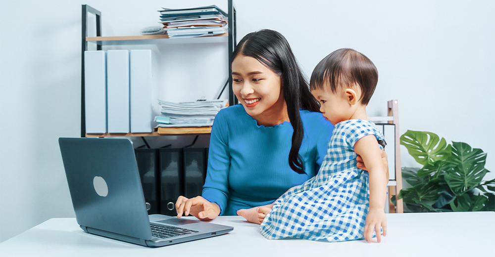 Mom working on a laptop at home while caring for her child, representing a flexible and family-friendly workplace.