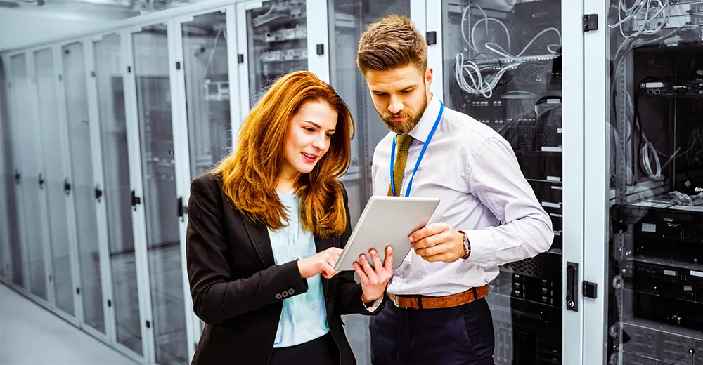 Two network systems administrators reviewing data on a tablet while working inside a server room, managing network system.