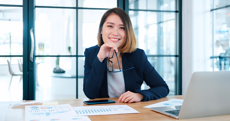Smiling accounting diploma graduate with glasses and reports at her desk.