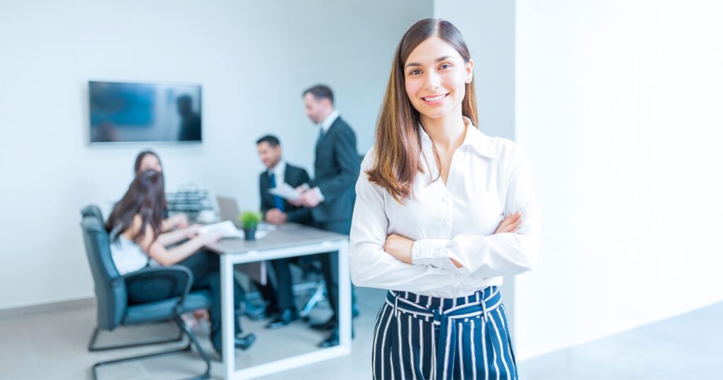 Female Business Administration Management Graduate ready for diverse responsibilities in a business setting. Her arms are crossed and a group of colleagues are in the background.