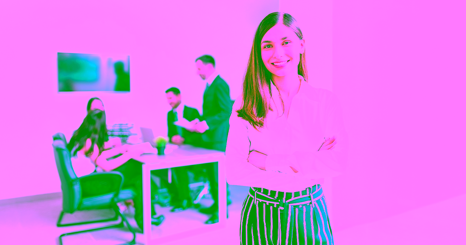 Female Business Administration Management Graduate ready for diverse responsibilities in a business setting. Her arms are crossed and a group of colleagues are in the background.