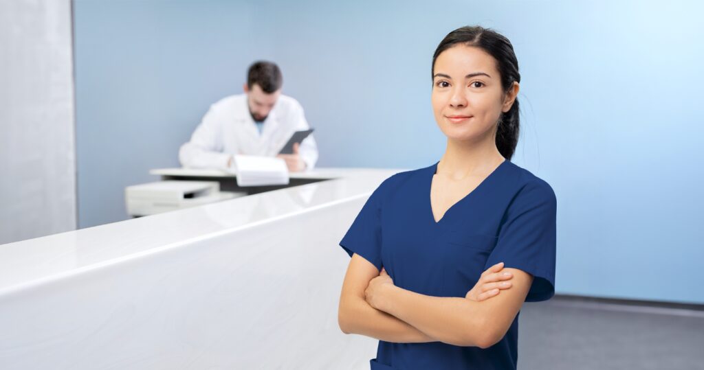 Female medical office administrator standing confidently in a healthcare setting with a doctor in the background.