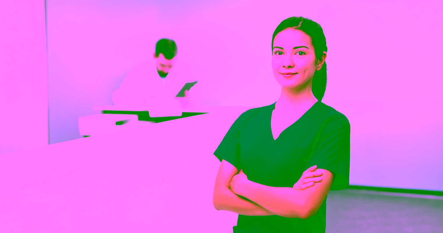 Female medical office administrator standing confidently in a healthcare setting with a doctor in the background.